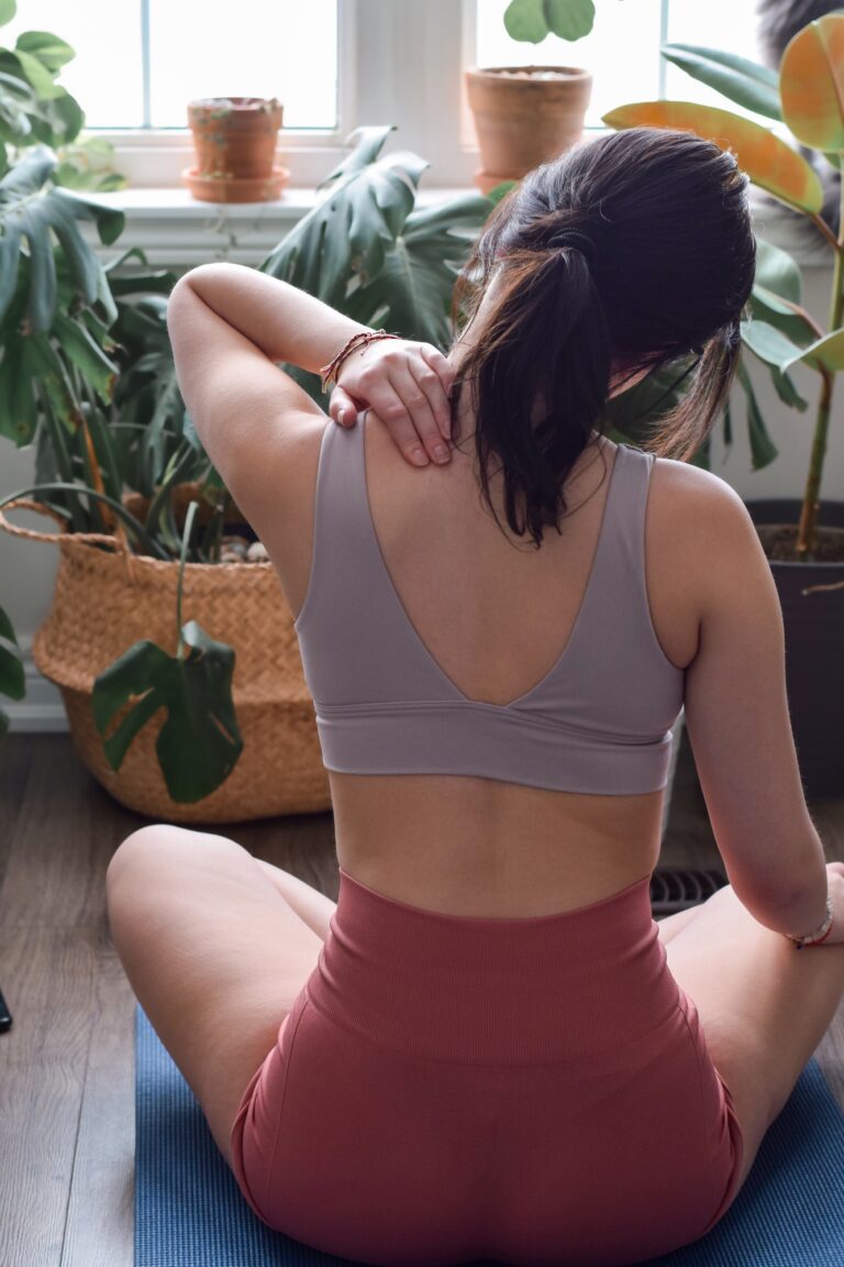 Young woman from behind sitting on yoga mat touching stretching sore neck muscles during workout